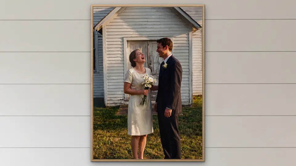 A framed poster of a laughing bride and groom outside a white country church, hanging on a white shiplap wall.